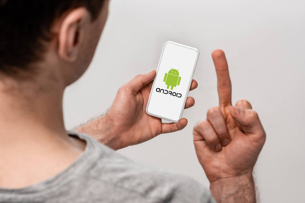 KYIV, UKRAINE - MAY 16, 2019: cropped view of man using smartphone with android app and showing middle finger, isolated on grey - 写真・画像