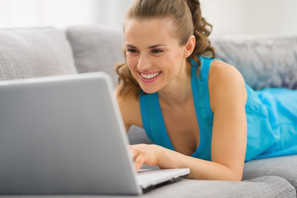 Smiling young woman laying on couch with laptop - Fotó, kép