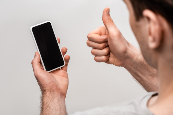 partial view of man showing thumb up while holding smartphone with blank screen isolated on grey - 写真・画像