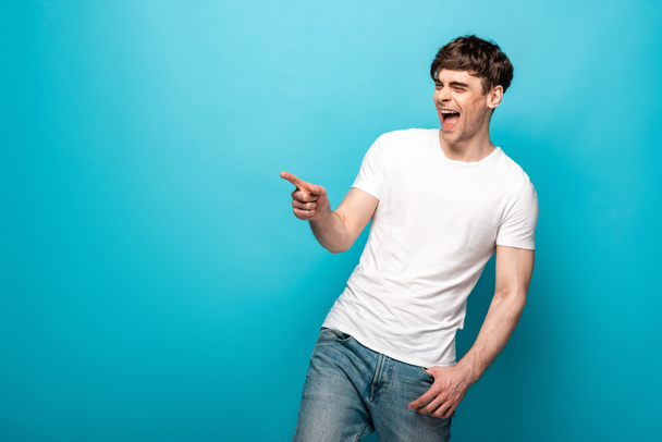 laughing man pointing with finger while looking away on blue background - Photo, Image