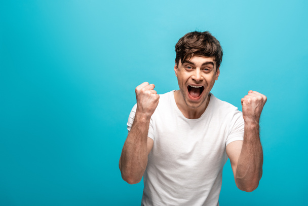 happy young man showing winner gesture while looking at camera on blue background - Фото, зображення