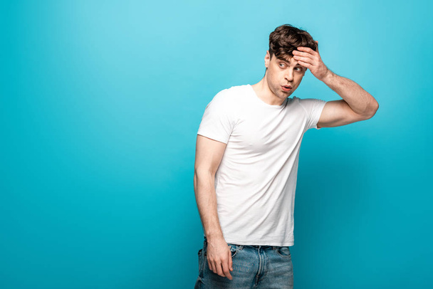 shocked young man looking away while holding hand on head on blue background - Фото, зображення