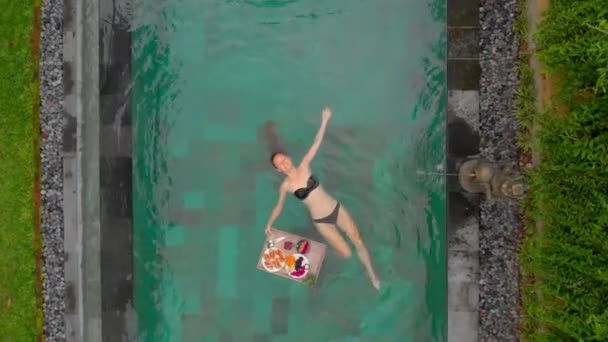 Aerial shot of a young woman tourist has her own personal breakfast on a floating table in a private swimming pool - Záběry, video