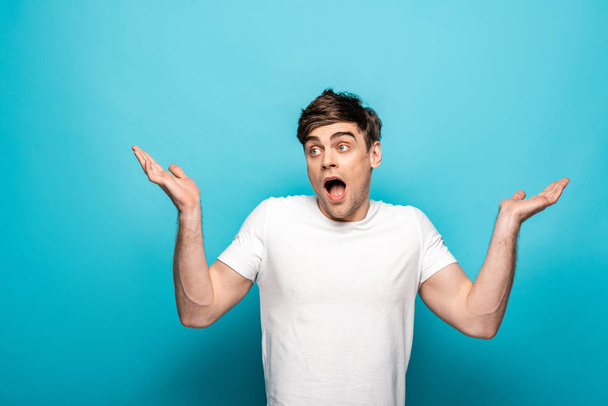 discouraged young man showing shrug gesture and looking away on blue background - Foto, imagen