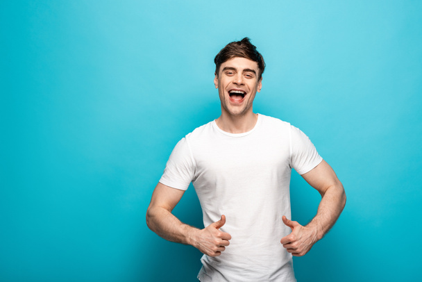 positieve jonge man glimlachend op de camera en toont duimen omhoog op blauwe achtergrond  - Foto, afbeelding