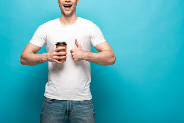 cropped view of smiling man holding coffee to go and showing thumb up on blue background - Fotoğraf, Görsel