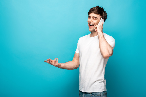 cheerful young man gesturing while talking on smartphone and looking away on blue background - Foto, afbeelding
