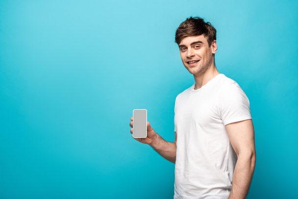 handsome young man showing smartphone with blank screen and smiling at camera on blue background - Foto, Bild