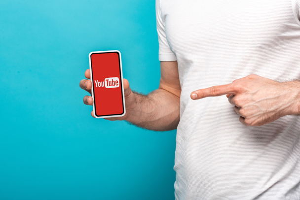 KYIV, UKRAINE - MAY 16, 2019: cropped view of man pointing at smartphone with youtube app, isolated on blue - 写真・画像