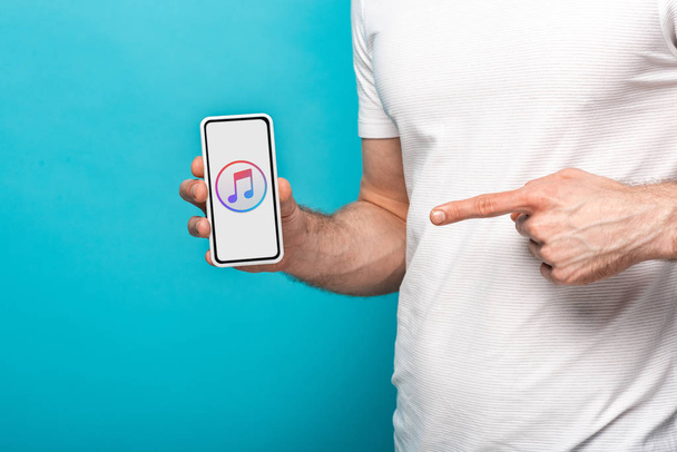 KYIV, UKRAINE - MAY 16, 2019: cropped view of man pointing at smartphone with apple music app, isolated on blue - Foto, imagen