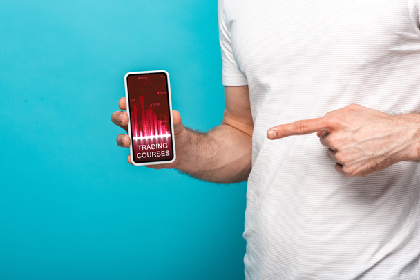 visão cortada do homem apontando para smartphone com aplicativo de cursos de negociação, isolado em azul
 - Foto, Imagem
