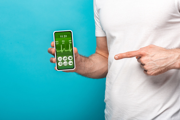 visão cortada do homem apontando para o smartphone com aplicativo de saúde, isolado no azul
 - Foto, Imagem