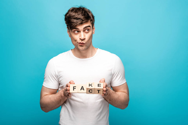 discouraged young man holding wooden cubes with fake fact lettering on blue background - Photo, Image