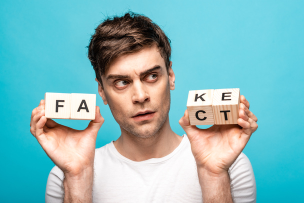 thoughtful young man looking away while holding wooden cubes with fake fact lettering isolated on blue - Photo, Image