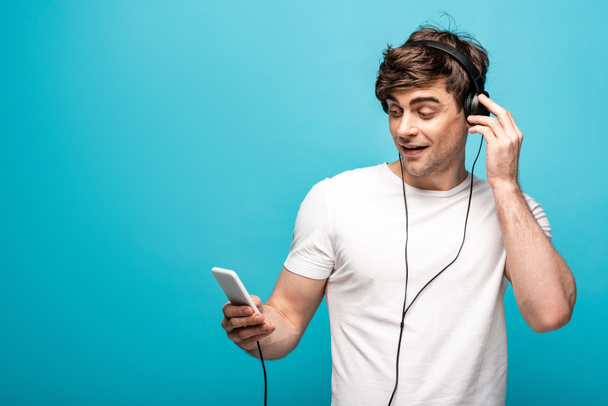 hombre guapo escuchando música en los auriculares y el uso de teléfono inteligente sobre fondo azul
 - Foto, Imagen