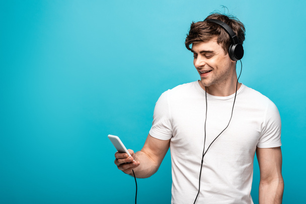 joven guapo sonriendo mientras escucha música con auriculares y teléfono inteligente sobre fondo azul
 - Foto, Imagen