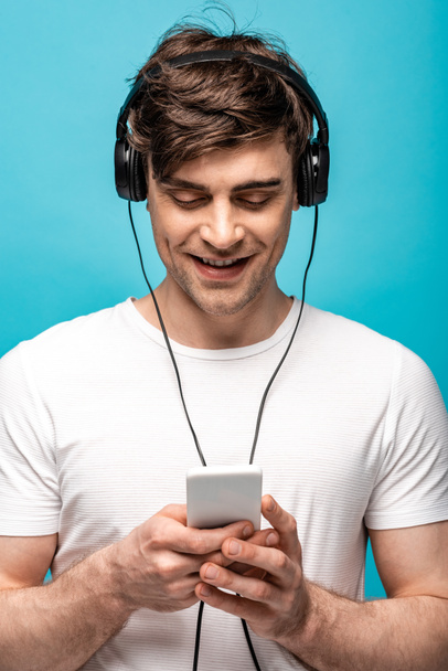 cheerful young man listening music in headphones while using smartphone isolated on blue - Foto, Bild