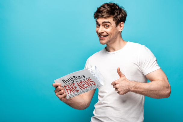 cheerful man showing thumb up while holding newspaper with fake news on blue background - Photo, Image