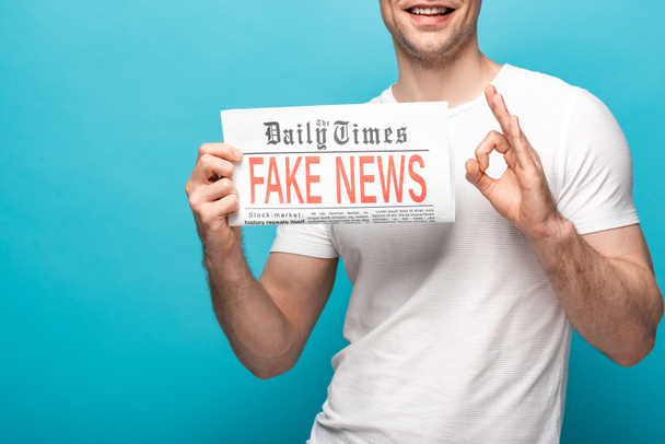 recortado vista de sonriente hombre mostrando ok signo mientras sostiene periódico con noticias falsas sobre fondo azul
 - Foto, Imagen