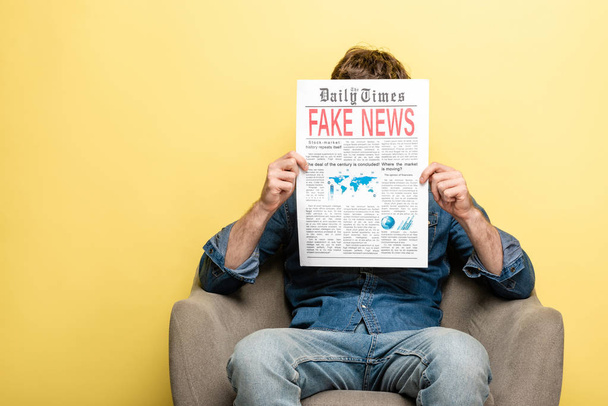 アームチェアに座り、黄色の背景に偽のニュースでニュースを読む男 - 写真・画像