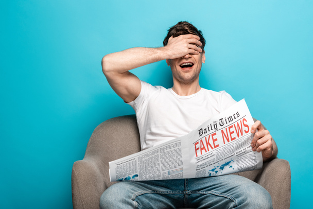 young man laughing while sitting in armchair and holding newspaper with fake news on blue background - Foto, Imagen