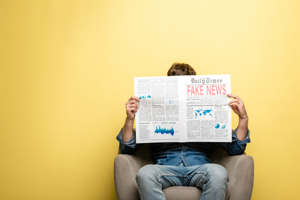 young man sitting in armchair and reading newspaper with fake news on yellow background - Foto, Bild