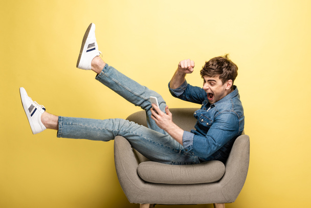izgatott fiatalember mutatja igen gesztus ülve fotel és használ smartphone sárga háttér - Fotó, kép