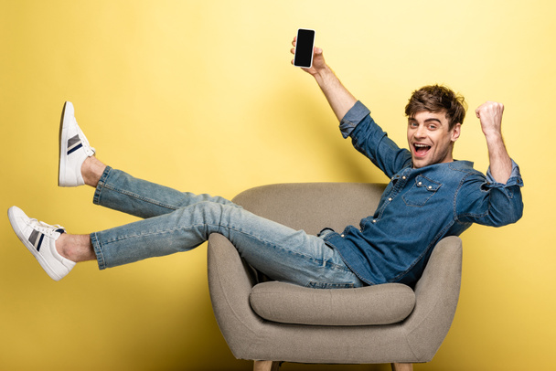 excited young man showing winner gesture while sitting in armchair and holding smartphone with blank screen on yellow background - Zdjęcie, obraz