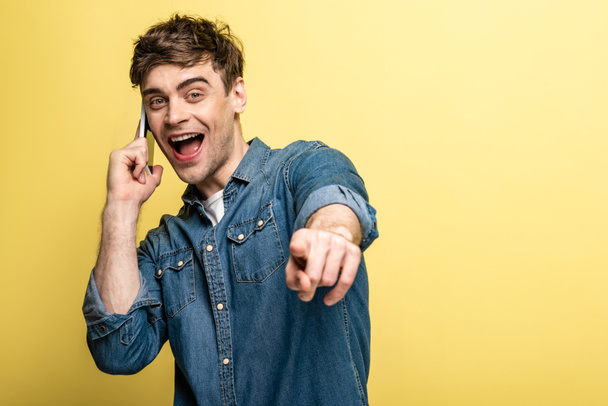excited young man pointing with finger at camera while talking on smartphone on yellow background - Fotó, kép