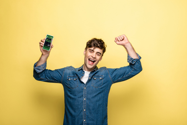 handsome smiling man in denim clothes holding smartphone with booking app on yellow - Photo, Image