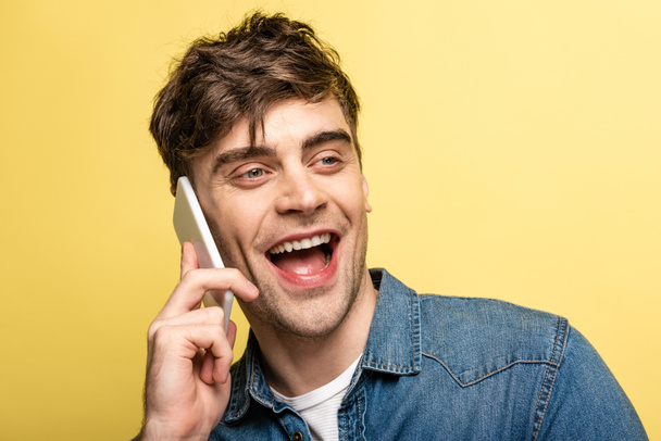 homem alegre olhando para longe e rindo enquanto fala no smartphone isolado no amarelo
 - Foto, Imagem