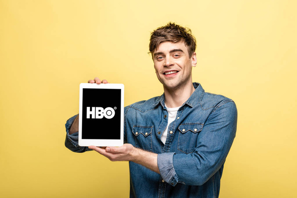 KYIV, UKRAINE - MAY 16, 2019: handsome cheerful man in jeans clothes showing digital tablet with HBO app, isolated on yellow - Φωτογραφία, εικόνα