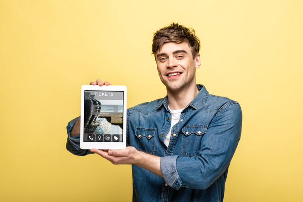 handsome smiling man in denim clothes showing digital tablet with tickets app, isolated on yellow - 写真・画像