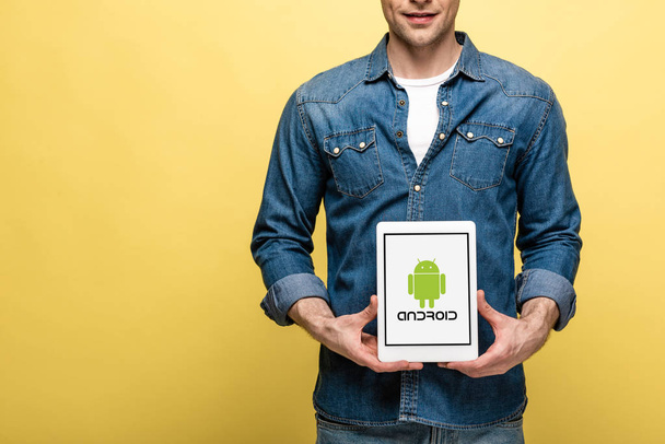 KYIV, UKRAINE - MAY 16, 2019: cropped view of man in jeans clothes showing digital tablet with android app, isolated on yellow - Fotoğraf, Görsel