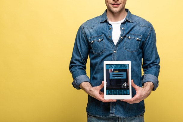 cropped view of man in denim clothes holding digital tablet with booking app, isolated on yellow - Photo, Image
