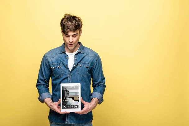 hombre guapo en ropa de mezclilla mirando tableta digital con tickets aplicación, aislado en amarillo
 - Foto, Imagen
