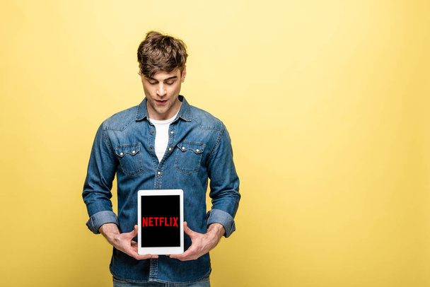 KYIV, UKRAINE - MAY 16, 2019: handsome man in jeans clothes looking at digital tablet with netflix app, isolated on yellow - Photo, Image