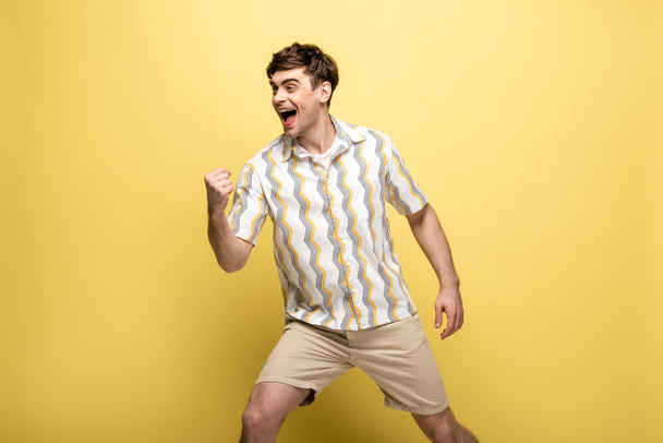 cheerful young man looking away and showing winner gesture on yellow background - Fotografie, Obrázek
