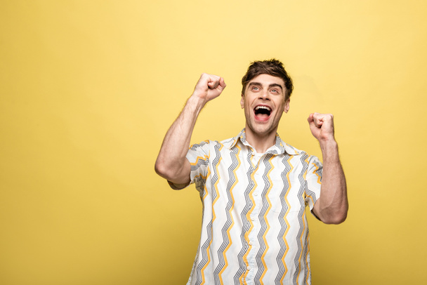 cheerful young man looking up and showing winner gesture on yellow background - Valokuva, kuva