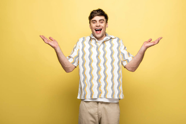 cheerful young man showing shrug gesture and smiling at camera on yellow background - Foto, afbeelding