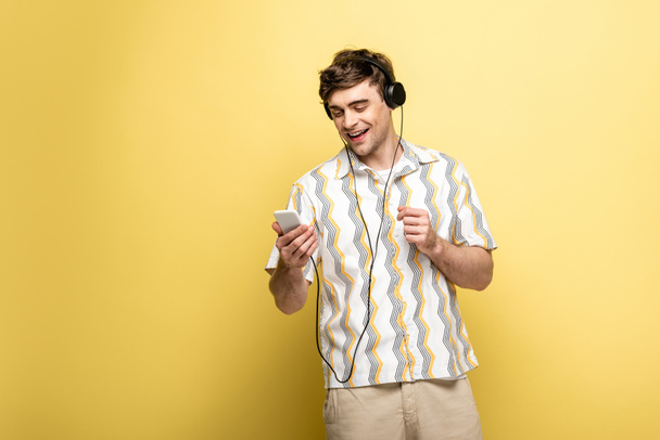 веселый молодой человек, слушающий музыку в наушниках и использующий смартфон на желтом фоне
 - Фото, изображение