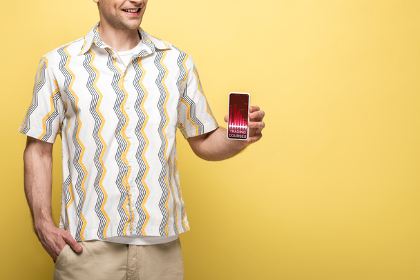 トレーディングコースでスマートフォンを見せる笑顔の男のトリミングビュー, 黄色に隔離 - 写真・画像