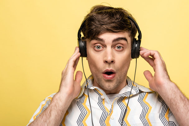 потрясенный молодой человек смотрит в камеру, слушая музыку в наушниках, изолированных на желтом
 - Фото, изображение