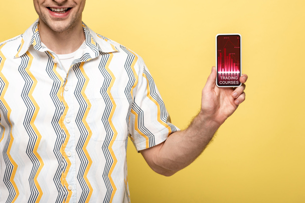 oříznutý pohled na usmívající se muž zobrazující smartphone s obchodními kurzy, izolované na žluté - Fotografie, Obrázek