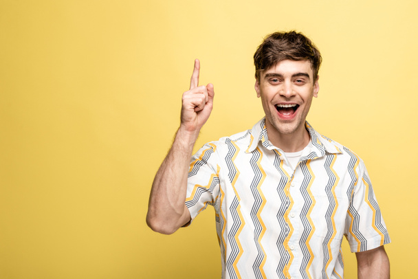 cheerful young man showing idea sign while smiling at camera on yellow background - Фото, изображение