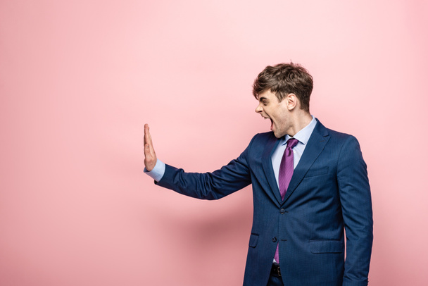 irritated businessman showing stop gesture while looking away on pink background - Zdjęcie, obraz