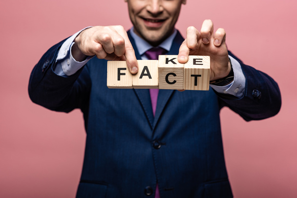 partial view of businessman holding wooden cubes with fake fact lettering on pink background - Photo, Image