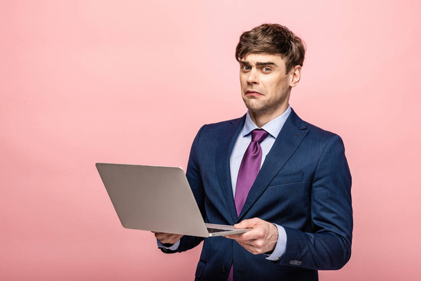 disappointed businessman looking at camera while holding laptop on pink background - Фото, зображення
