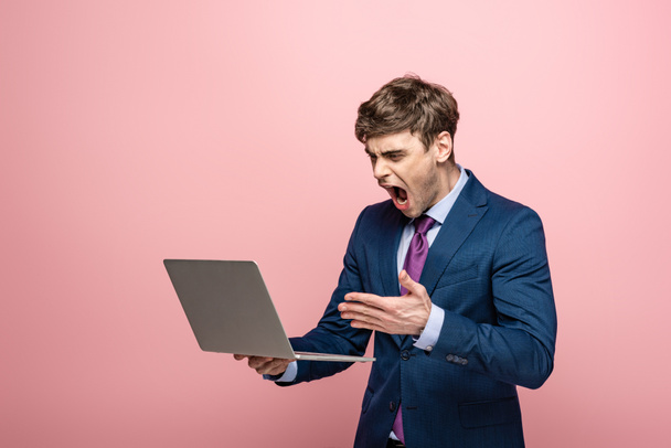 angry businessman quarreling while looking at laptop isolated on pink - Фото, зображення
