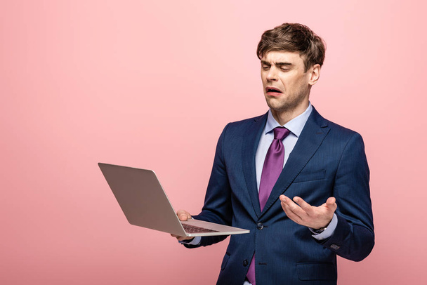 displeased businessman gesturing while looking at laptop on pink background - Fotoğraf, Görsel